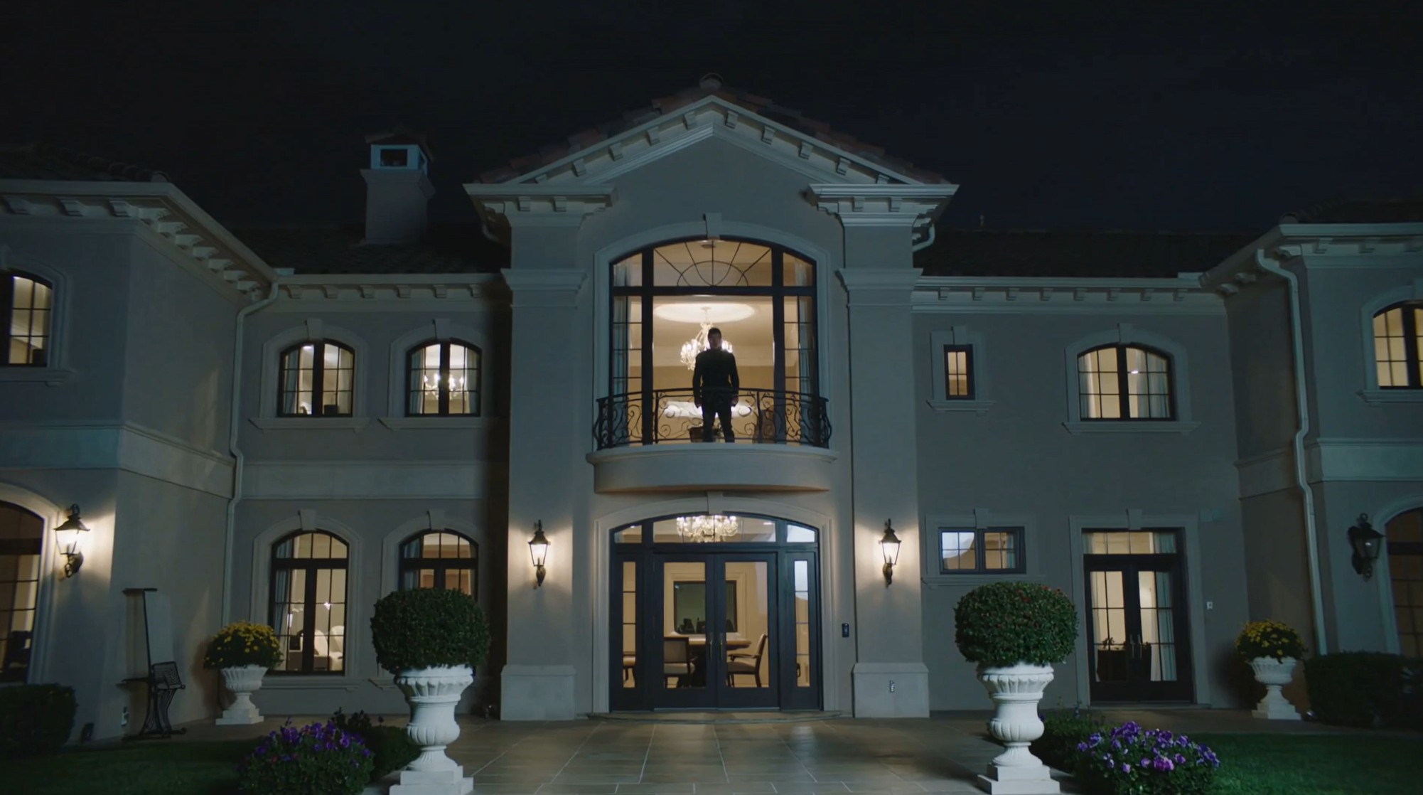 A solitary figure stands on the lit balcony of an imposing country club at night