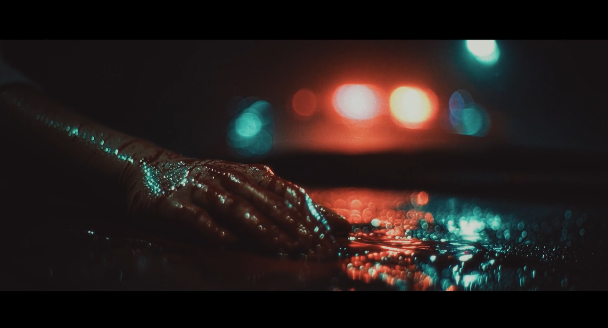 A bloodied hand lies on wet pavement beneath blurred emergency lights